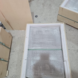 Puede incluir: Un objeto rectangular envuelto en pl&aacute;stico de burbujas transparente y asegurado con una cinta de pl&aacute;stico verde, colocado dentro de una caja de madera clara forrada con espuma blanca. La caja est&aacute; abierta por arriba, mostrando el embalaje.