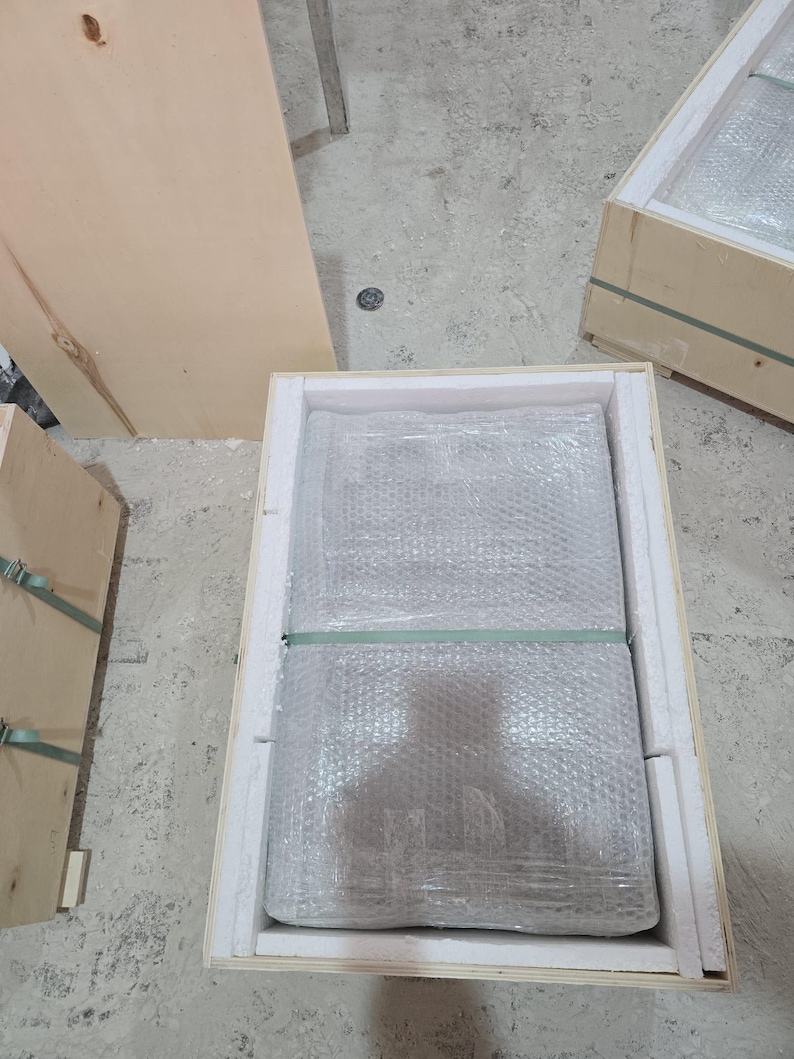 Puede incluir: Un objeto rectangular envuelto en pl&aacute;stico de burbujas transparente y asegurado con una cinta de pl&aacute;stico verde, colocado dentro de una caja de madera clara forrada con espuma blanca. La caja est&aacute; abierta por arriba, mostrando el embalaje.