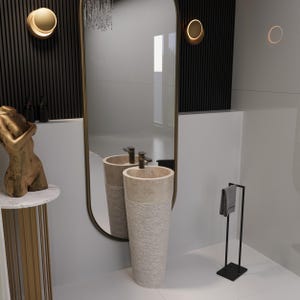 May include: A modern bathroom scene featuring a tall, textured stone pedestal sink with a matching countertop. A large, arched mirror reflects the sink and surrounding decor, including a gold-toned sculpture and sconces. A black towel rack holds a gray towel.