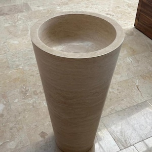 May include: A tall, cylindrical stone sink with a wide, circular basin. The sink is made of a light beige stone with subtle veining, and it stands on a tiled floor. The design is modern and minimalist.