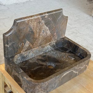 May include: A brown marble sink with a rectangular basin and a decorative backsplash. The sink has a natural stone pattern with shades of brown, gray, and white. The sink is placed on a wooden pallet.
