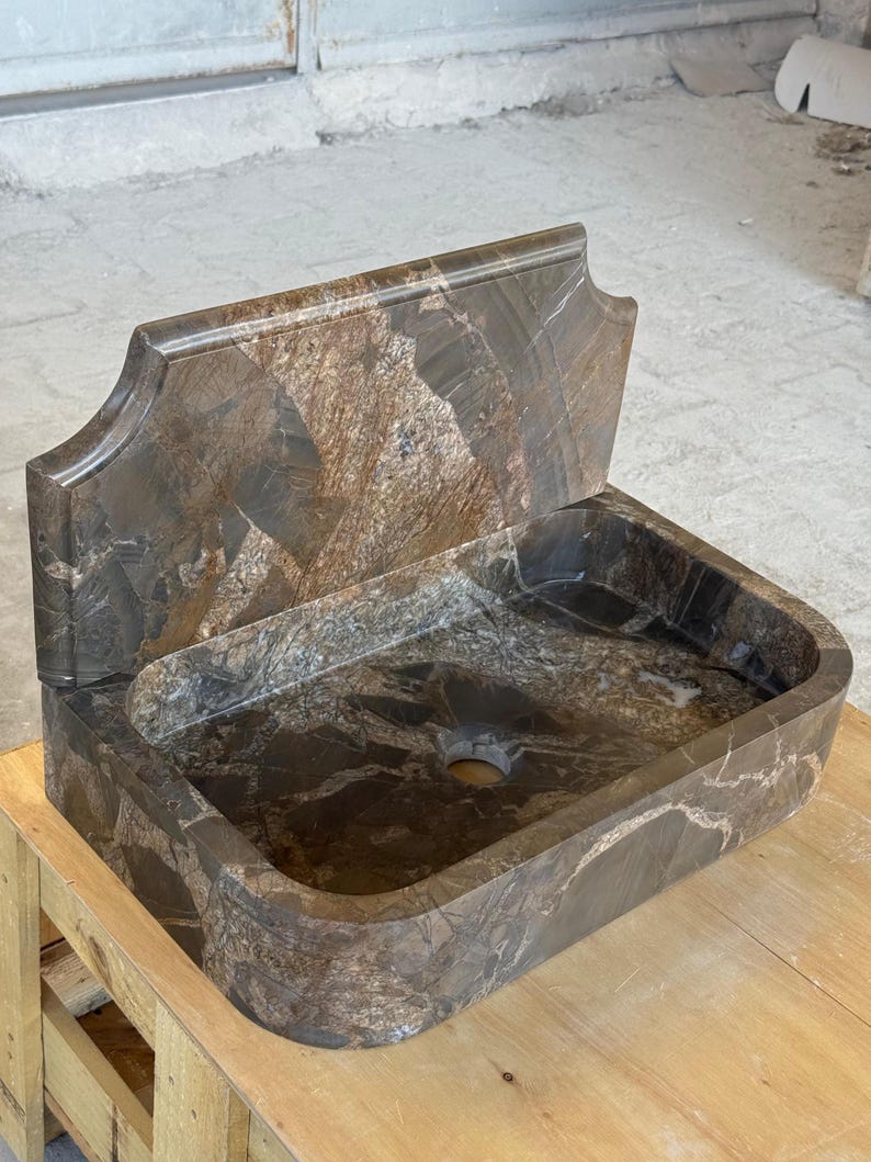 May include: A brown marble sink with a rectangular basin and a decorative backsplash. The sink has a natural stone pattern with shades of brown, gray, and white. The sink is placed on a wooden pallet.