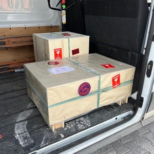 Puede incluir: Dos cajas de env&iacute;o rectangulares de madera envueltas en papel color canela claro, aseguradas con correas de pl&aacute;stico verde. Las cajas tienen etiquetas rojas de fr&aacute;gil con un icono de copa de vino, un sello circular morado y etiquetas de env&iacute;o.
