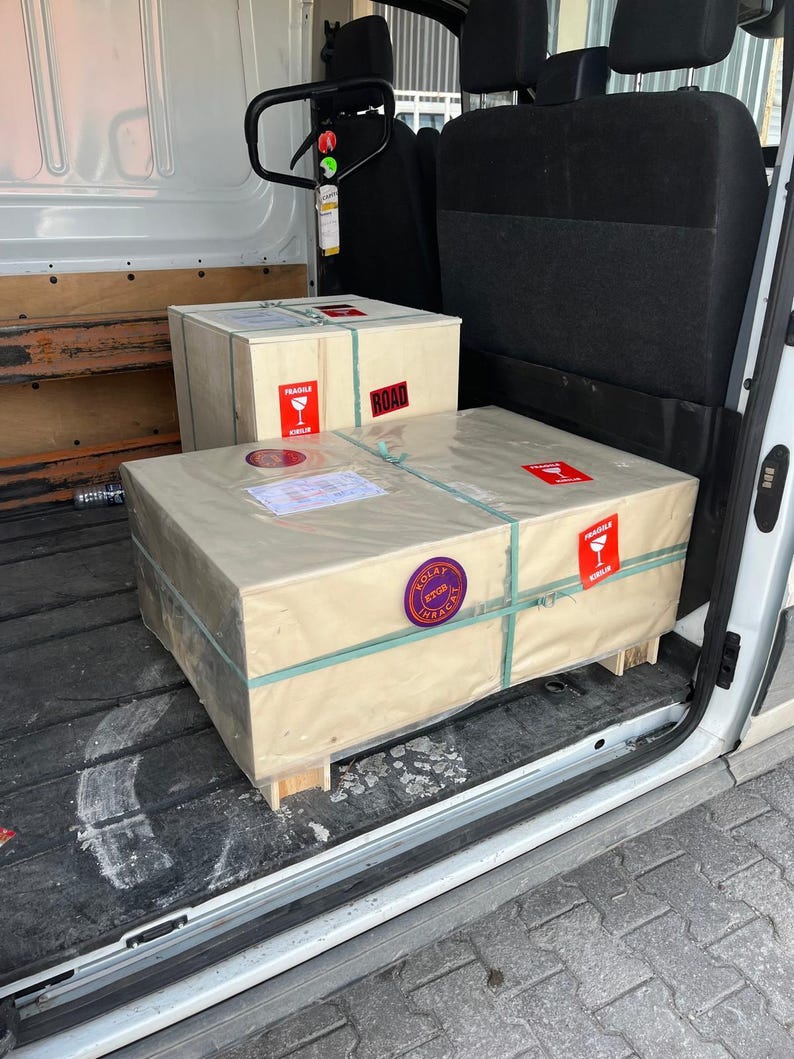 Puede incluir: Dos cajas de env&iacute;o rectangulares de madera envueltas en papel color canela claro, aseguradas con correas de pl&aacute;stico verde. Las cajas tienen etiquetas rojas de fr&aacute;gil con un icono de copa de vino, un sello circular morado y etiquetas de env&iacute;o.