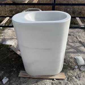May include: A large, white, freestanding sink with a rounded rectangular shape. The sink has a wide, oval basin and a smooth, polished surface. It is made of a solid, opaque material and sits on a wooden base. The sink is outdoors.