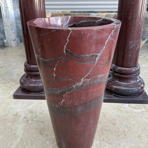 May include: A tall, tapered vase made of red marble with gray and white veining. The vase has a wide opening and a smooth, polished surface. Two matching marble columns are in the background.
