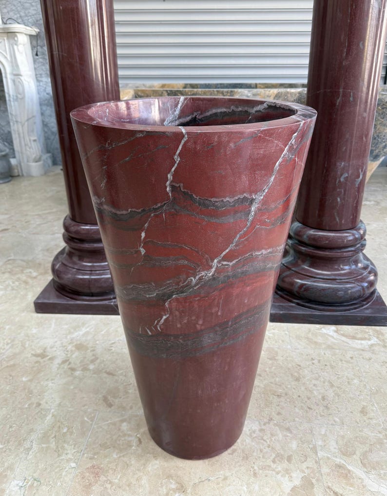 May include: A tall, tapered vase made of red marble with gray and white veining. The vase has a wide opening and a smooth, polished surface. Two matching marble columns are in the background.