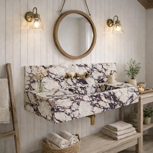 Puede incluir: Un lavabo de pared hecho de m&aacute;rmol blanco con vetas p&uacute;rpuras prominentes y grifos de lat&oacute;n. El lavabo se sit&uacute;a bajo un espejo redondo con marco de madera, flanqueado por dos apliques de pared de lat&oacute;n con pantallas de cristal.