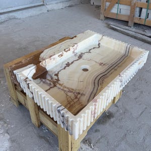 May include: A rectangular onyx sink with a light beige and brown marbled pattern. The sink has a decorative, ridged edge and is set on a wooden pallet. The sink features a single drain hole.