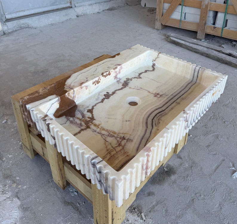 May include: A rectangular onyx sink with a light beige and brown marbled pattern. The sink has a decorative, ridged edge and is set on a wooden pallet. The sink features a single drain hole.