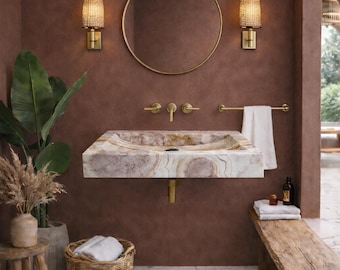 Puede incluir: Un lujoso cuarto de baño con un lavabo de mármol, grifos dorados y un espejo redondo. Las paredes son de un tono cálido y terroso. Un banco de madera, una cesta de mimbre y plantas en macetas completan el ambiente. Apliques iluminan la estancia.