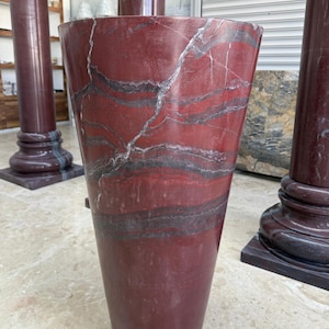 May include: A tall, tapered vase made of red marble with dark gray and white veining. The vase has a wide top and a narrower base. The marble has a polished finish. Other marble columns are visible in the background.