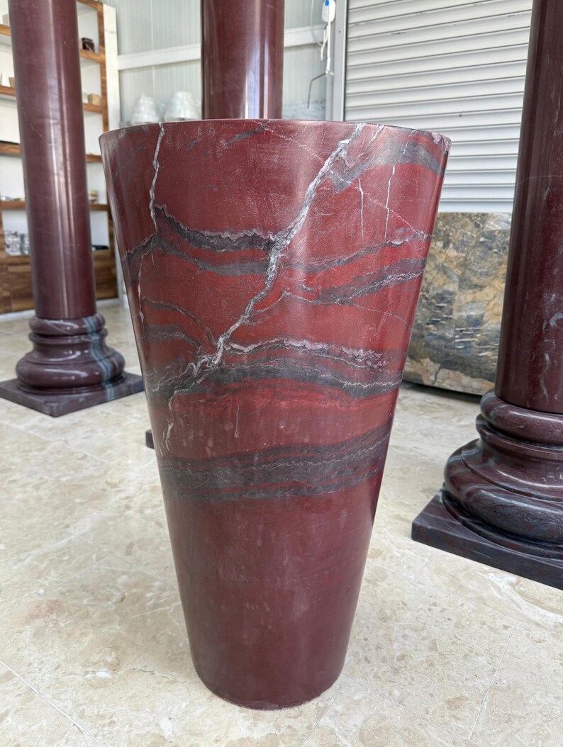 May include: A tall, tapered vase made of red marble with dark gray and white veining. The vase has a wide top and a narrower base. The marble has a polished finish. Other marble columns are visible in the background.