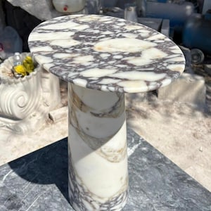 Può includere: Un tavolino in marmo con piano rotondo e base cilindrica. Il tavolo presenta uno sfondo bianco con venature grigie e marroni. Il tavolo è in pietra ed è adatto per uso interno o esterno.