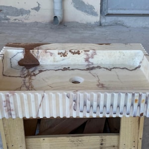 May include: Rectangular marble sink with a light beige and brown color scheme. The sink has a textured, ribbed edge and a central drain hole. The sink is placed on a wooden pallet.