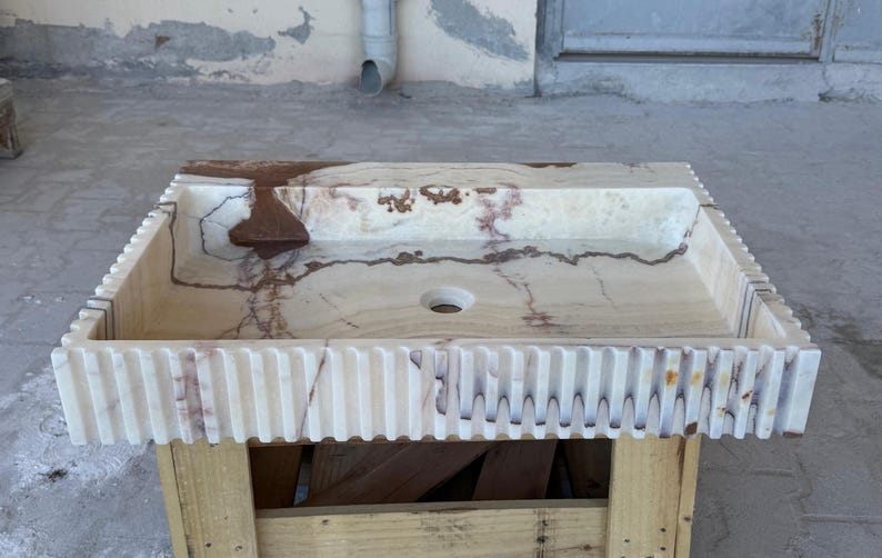 May include: Rectangular marble sink with a light beige and brown color scheme. The sink has a textured, ribbed edge and a central drain hole. The sink is placed on a wooden pallet.