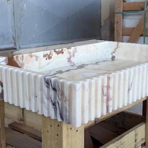 May include: Rectangular onyx sink with a fluted exterior. The sink features a cream and brown marbled pattern. It is displayed on a wooden pallet. The sink's dimensions are not visible.