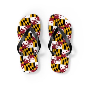 Może przedstawiać: Para klapek z wzorem flagi stanu Maryland. Sandały mają czarny pasek i białe tło z geometrycznym wzorem flagi stanu w kolorze czerwonym, czarnym, żółtym i białym. Klapki są pokazane na białej powierzchni.