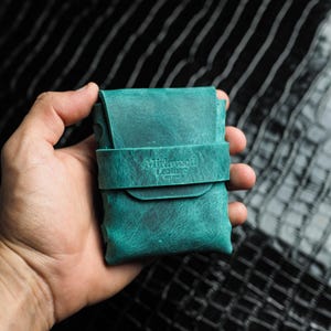 Peut inclure: Un portefeuille en cuir de couleur teal avec une fermeture à rabat et une sangle. La sangle porte l'inscription "Midwood Leather". Le portefeuille est tenu dans une main sur un fond rayé noir et blanc.