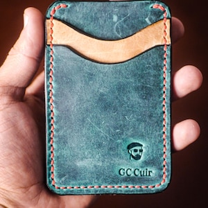 Peut inclure: Un porte-cartes en cuir bleu-vert avec une poche marron clair et des coutures rouges. Le porte-cartes porte le logo "GC Cuir" en relief dans le coin inférieur droit. Le cuir a un aspect vieilli.