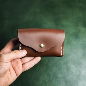 Peut inclure: Un porte-cartes en cuir marron avec un rabat incurvé fermé par un bouton-pression en métal couleur laiton. Cet accessoire rectangulaire présente des coutures blanches visibles sur les bords latéraux, offrant un design épuré et fonctionnel.