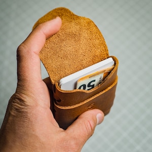 Peut inclure: Un porte-cartes en cuir marron, ouvert pour révéler des cartes blanches et un billet de 50 euros. Le porte-cartes est en cuir texturé avec des coutures visibles. Le rabat est ouvert.