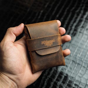Peut inclure: Un portefeuille en cuir marron avec une fermeture à rabat et une sangle. Le portefeuille porte les mots "Midwest Leather" en relief sur la sangle. Le portefeuille est tenu dans une main.