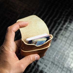 Peut inclure: Un porte-cartes en cuir marron clair, ouvert pour révéler plusieurs cartes et de l'argent liquide. Le porte-cartes est tenu dans une main, mettant en valeur son design compact et la texture naturelle du cuir. Le mot "Midwood" est visible en bas.