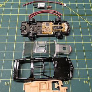 以下が含まれることがあります： 分解された黒いおもちゃの車。車輪、回路基板、配線など、内部コンポーネントが見えます。部品は緑色のカッティングマット上に配置されており、車の構造と電子機器を示しています。