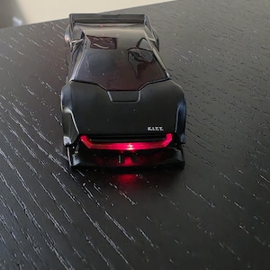 Puede incluir: Un modelo de coche de juguete negro con un diseño elegante y angular, que presenta un parabrisas tintado oscuro y una prominente barra de luz roja en la parrilla delantera. Las letras K.I.T.T. están impresas en blanco en el capó delantero.