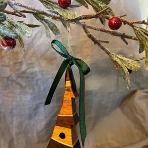 May include: A wooden Christmas tree ornament in the shape of a tree, with a dark green ribbon. The ornament is made from various shades of wood, including light and dark brown, and features a small, round hole. It hangs from a gold wire, set against frosted greenery and red berries.