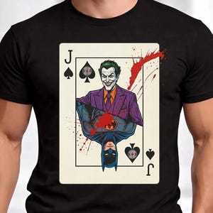 May include: Black t-shirt featuring a playing card design with the Joker and Batman. The Joker is depicted in a purple suit, and Batman is shown upside down. The card is a Jack of Spades with red splatter accents.