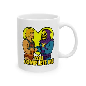 May include: White ceramic mug featuring a cartoon image of He-Man and Skeletor holding hands inside a yellow heart. The text "YOU COMPLETE ME" is below the characters. The mug has a curved handle.