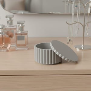 May include: A light gray, ribbed, round trinket box with its lid slightly ajar, displayed on a light wood surface. Several perfume bottles and a jewelry stand with necklaces and earrings are also visible, suggesting a vanity or dressing table setting.