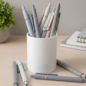 May include: A collection of pastel-colored pens in a white cylindrical holder. The pens are in shades of blue, gray, and pink, with gold-colored accents. Several pens are scattered on the light-colored wooden surface.