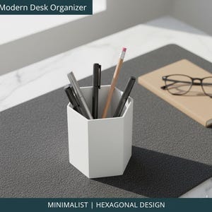 May include: A white hexagonal desk organizer holding pens and a pencil. A notebook and glasses sit on a gray surface. The text "Modern Desk Organizer" and "MINIMALIST | HEXAGONAL DESIGN" are visible.
