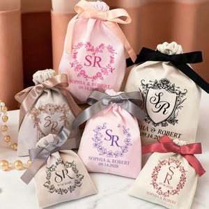 May include: Collection of drawstring favour bags in shades of pink, cream, and beige. Each bag is adorned with a floral design, the initials "SR", the names "SOPHIA & ROBERT", and a date. The bags are finished with satin ribbons in coordinating colours.