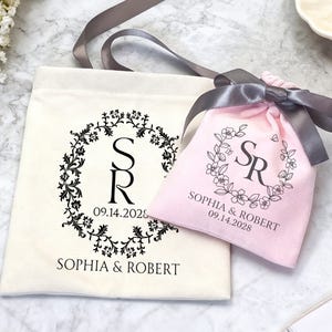 May include: Two personalized wedding favor bags. One is a cream-colored tote bag with a black floral wreath design and the initials "SR" and the date "09.14.2028". The other is a pink drawstring bag with a gray ribbon and the same design and text.