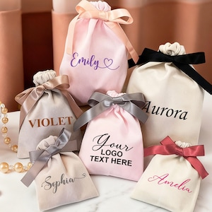 Könnte beinhalten: Eine Sammlung von Geschenkbeuteln mit Kordelzug in verschiedenen Farben, darunter Rosa, Beige und Weiß. Jeder Beutel hat ein Satinband und personalisierten Text, wie Namen oder ein Logo. Die Beutel sind auf einer weißen Oberfläche angeordnet.