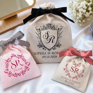 Puede incluir: Tres pequeñas bolsas con cordón y lazos decorativos. Una es rosa con un lazo gris, otra es blanquecina con un lazo negro y otra es blanquecina con un lazo rojo. Cada bolsa tiene un diseño floral con iniciales y una fecha. Las bolsas están sobre satén blanco.