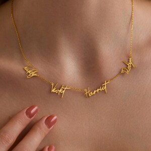 Op de afbeelding: Een goudkleurige ketting met de namen "Teddy", "Violet", "Harriet" en "Ruby" in een sierlijk handschrift. De ketting wordt om de nek gedragen. De namen zijn verbonden door een fijne ketting.