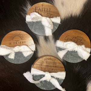 May include: Four round coasters with a wood top and a gray stone base. Each coaster has a white ribbon and a different inspirational quote engraved on the wood. The quotes include "Faith", "Be the Light", "God's Plan", and "Will Fear No".