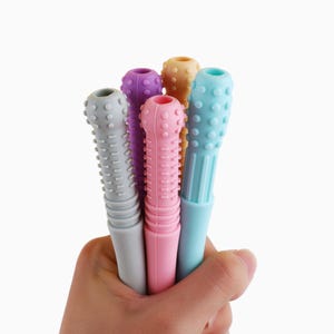 Könnte beinhalten: Eine Hand hält sechs bunte Baby-Beißspielzeuge. Die Spielzeuge sind in Grautönen, Lila, Rosa, Orange und Blau gehalten. Jedes Spielzeug hat eine strukturierte Oberfläche mit abgerundeten Noppen und Rillen, die zur Beruhigung wunden Zahnfleischs dienen.