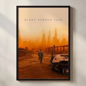 May include: Framed poster featuring the text "BLADE RUNNER 2049" above a dystopian cityscape. A figure walks away from a futuristic car in a desert landscape, with a towering city in the background, all in warm orange tones.