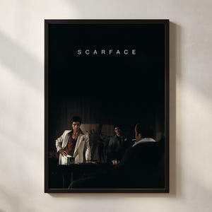 May include: A framed movie poster for Scarface featuring the title in white text at the top. The artwork depicts a scene with a person in a white suit standing in a dimly lit room, with other figures partially visible in the shadows nearby.