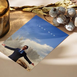 May include: A print featuring a man in a suit with arms outstretched against a blue sky background. The words "IRON MAN" are printed in white. The print is on a light-colored surface, with a brown glass bottle and decorative dried plants.
