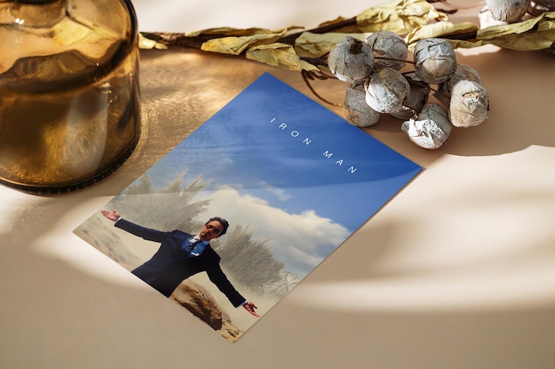 May include: A print featuring a man in a suit with arms outstretched against a blue sky background. The words "IRON MAN" are printed in white. The print is on a light-colored surface, with a brown glass bottle and decorative dried plants.