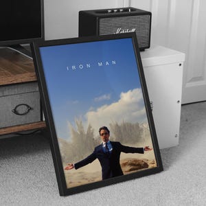 May include: Framed "Iron Man" movie poster featuring Robert Downey Jr. in a suit with arms outstretched against a blue sky and cloud backdrop. The title "IRON MAN" is displayed at the top. The poster is in a black frame.