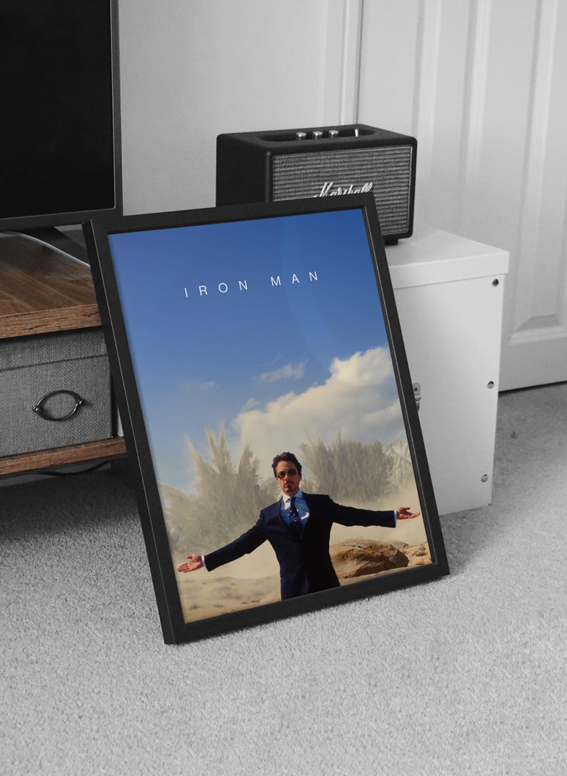 May include: Framed "Iron Man" movie poster featuring Robert Downey Jr. in a suit with arms outstretched against a blue sky and cloud backdrop. The title "IRON MAN" is displayed at the top. The poster is in a black frame.