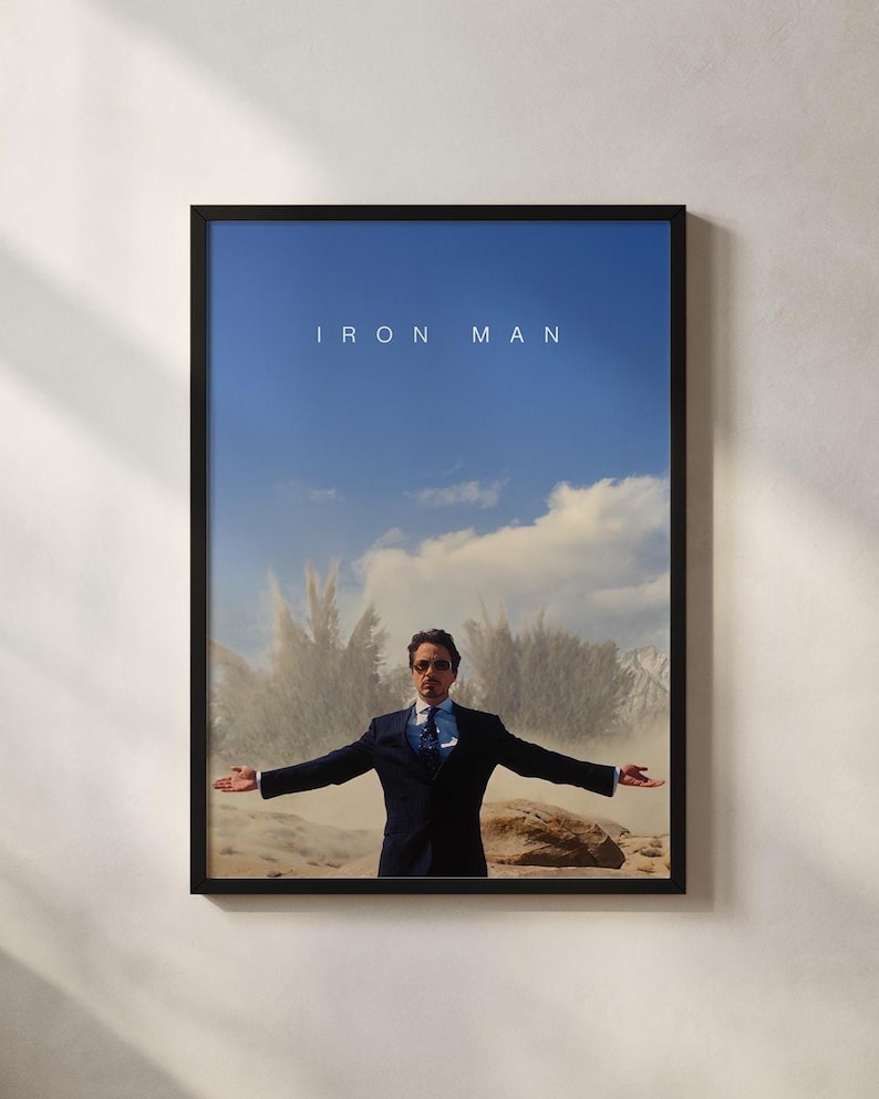 May include: Framed poster featuring a man in a suit with arms outstretched against a backdrop of a blue sky and a sand-colored landscape. The text "IRON MAN" is displayed at the top of the poster.
